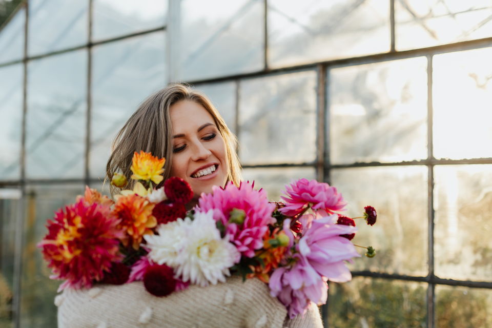 A florist laughing behind an armful of dahlias in a sunlit greenhouse.